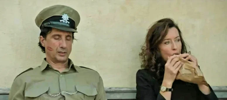 The policeman (Peter Galea) naps on the bench in front of the police station while Carmen (Natasha McElhone) has breakfast beside him - Carmen