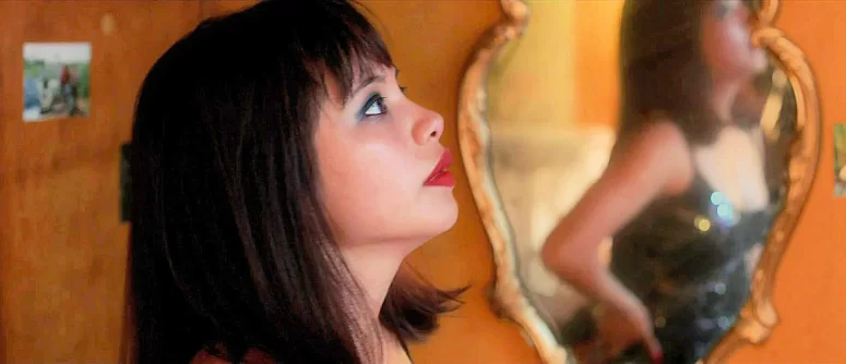 April communes with her reflection. On the wall behind is a photo from her past.